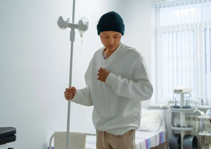 A person in a hospital room, wearing a beanie and an IV, walking with an IV stand, reflecting hard truths.