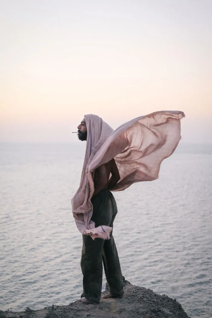 A man with a head covering standing on a cliff overlooking the Strait of Hormuz, with the fabric blowing in the wind.