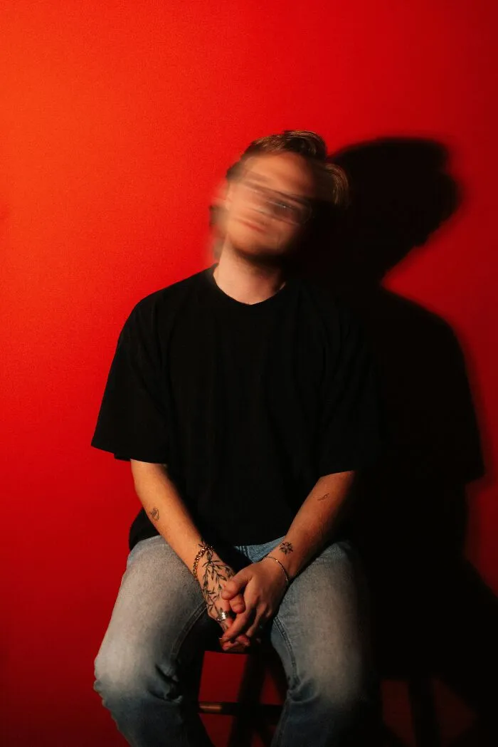 A young man sitting against a red wall with a blurred face, representing 'under my roof' rules that backfired on parents.