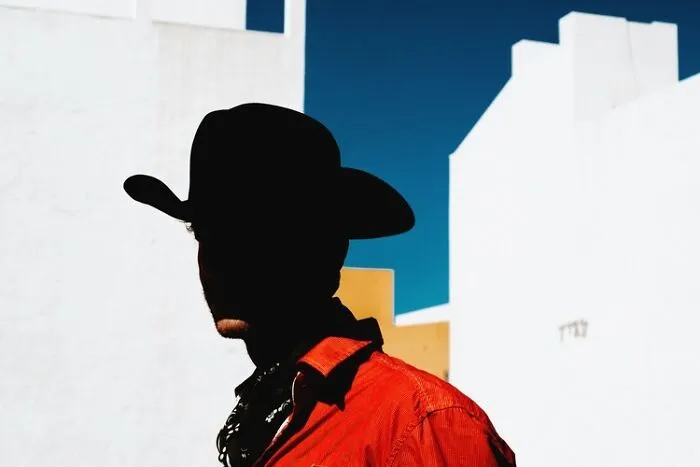 A person in a cowboy hat with a shadowed face and red shirt against white buildings, a remarkable color photo from the reFocus Contest.