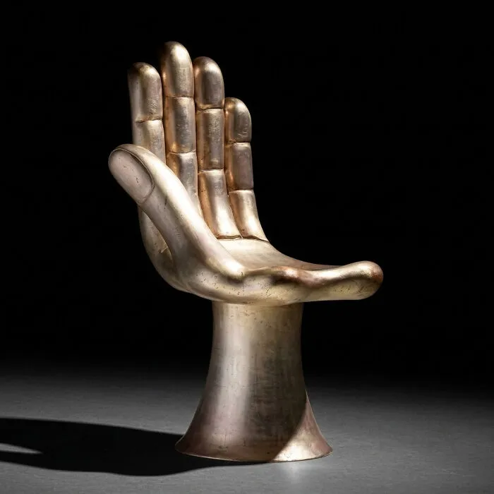 A gold-colored chair shaped like an open hand with fingers pointing upwards, a creation by furniture designers.