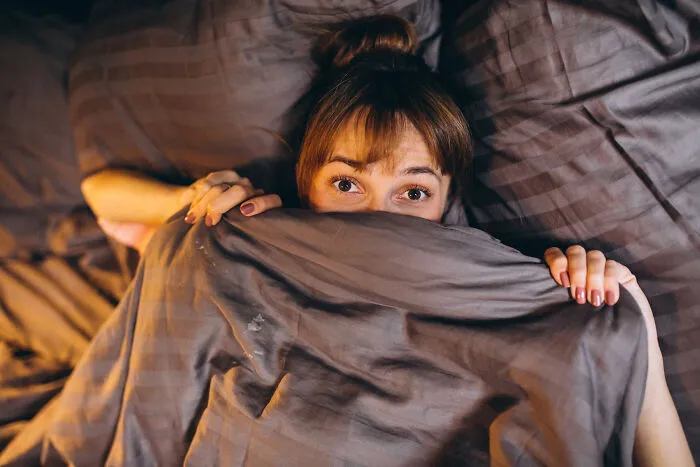 A woman peeking from under a duvet, looking scared, illustrating stories about guests who completely ignored house etiquette.