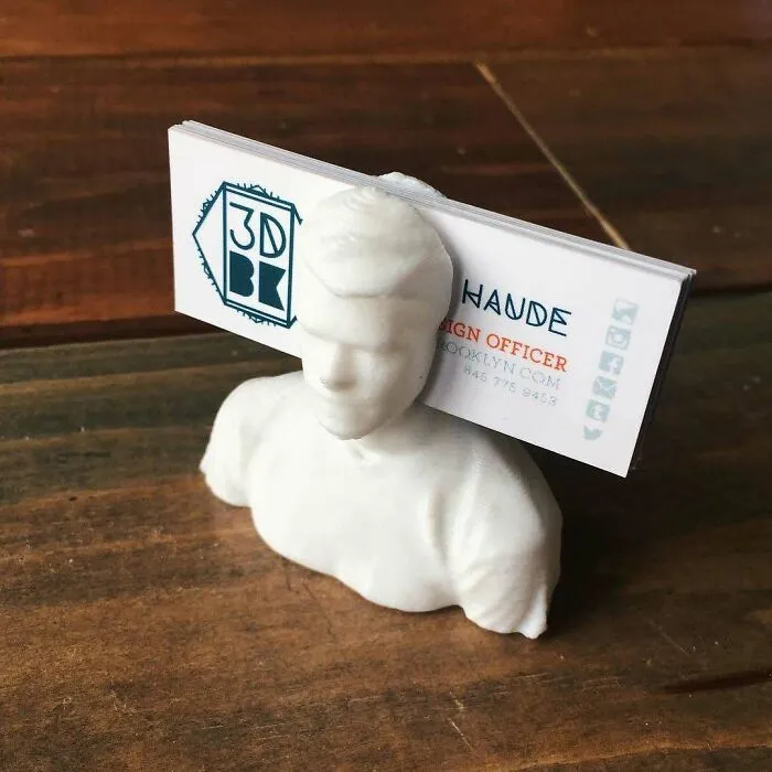 A white 3D printing creation of a bust holding business cards on a wooden surface. Amazing 3D printing creations.