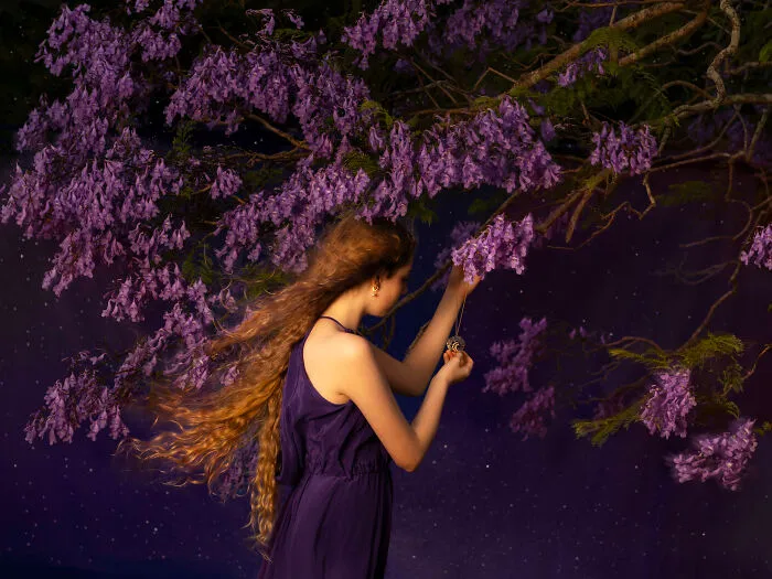 Surreal photo of a woman with long hair in a purple dress reaching for a branch with purple flowers against a starry background.