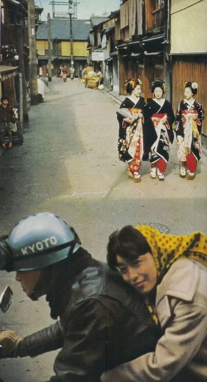 A historical photo of a street in Kyoto with a motorcycle rider and passengers, and three women in kimonos walking, a past scene.