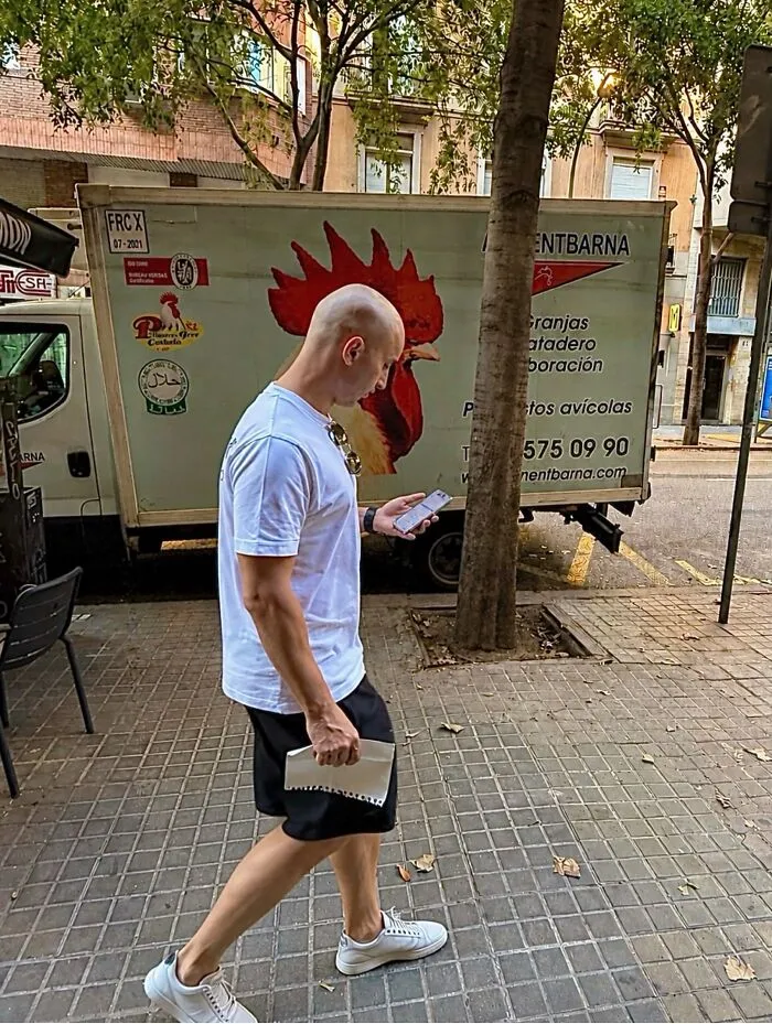 Un hombre con camiseta blanca y pantalones cortos negros caminando por la calle, con divertidas fotos accidentales al fondo.