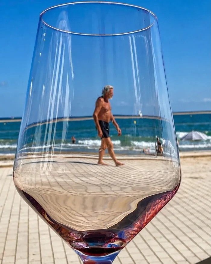 Fotos accidentales: hombre caminando en la playa, reflejado en una copa de vino, capturado en el momento justo.