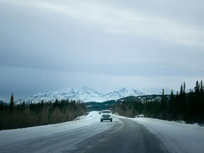 A vehicle drives on a snowy road with mountains in the background, a chilling internet rabbit hole.