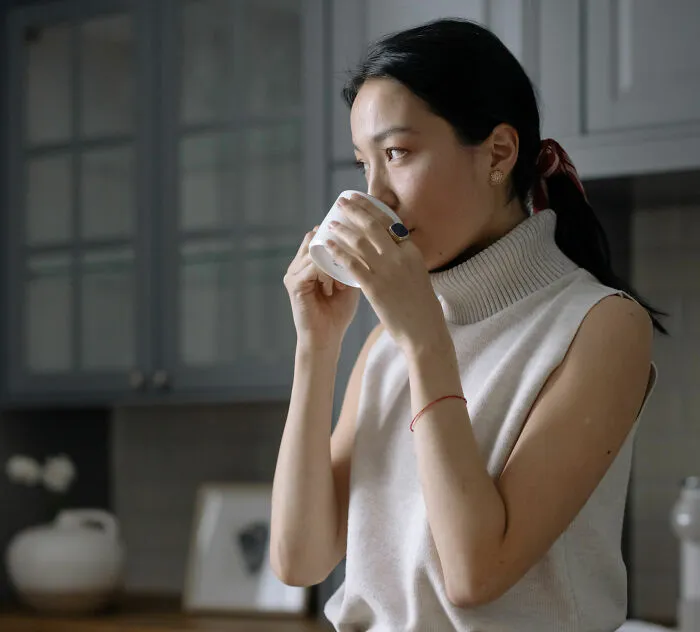 A woman in a cream turtleneck sips from a mug in a modern kitchen, reflecting an international couple's culture shock.