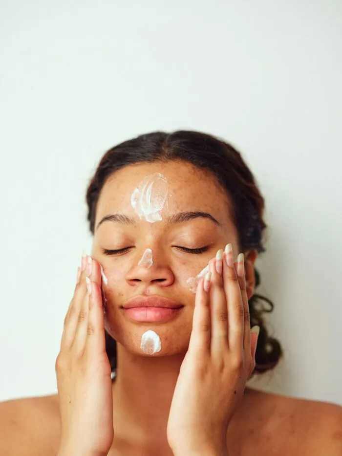 Woman applying face cream with dots on her forehead, nose, and chin, revealing companies' beauty secrets.