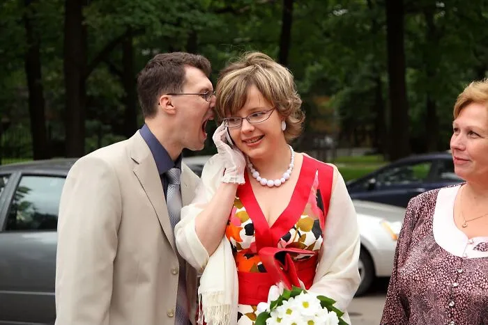 Funny wedding photo of a groom shouting into his bride's ear while she's on the phone.