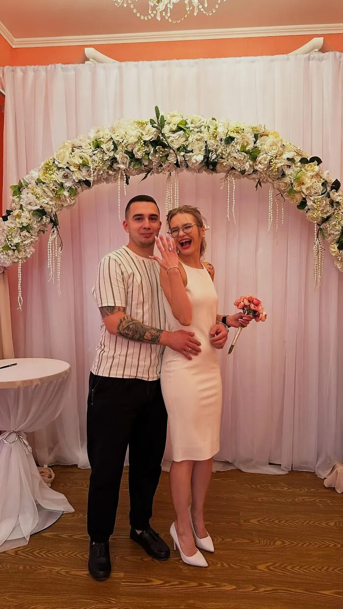 A happy couple showing off a ring under a floral arch, a funny wedding photo of their engagement joy.