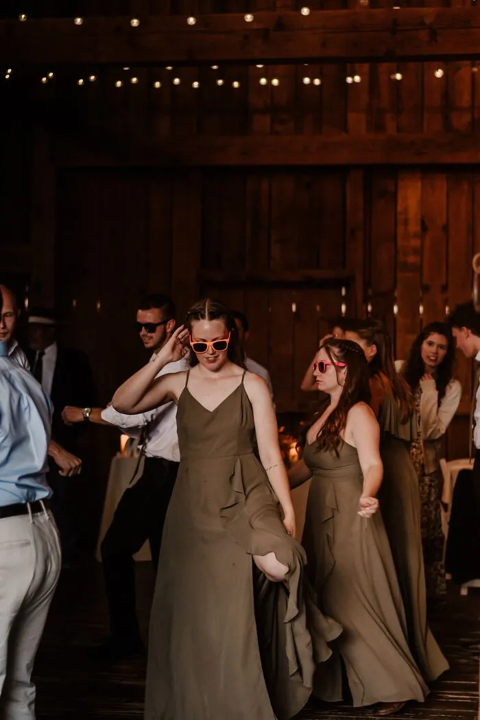 A funny wedding photo of bridesmaids wearing pink heart-shaped sunglasses and dancing at a rustic wedding reception.