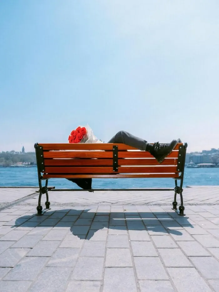 A person lounging on a park bench by the water, holding a bouquet of red roses, captured in a well-timed street shot.
