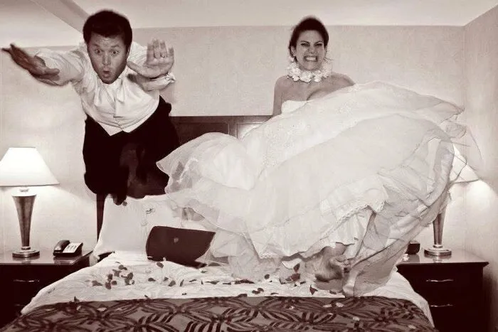 Iconic funny wedding photo of a newlywed couple mid-air jumping on a bed in their wedding attire.