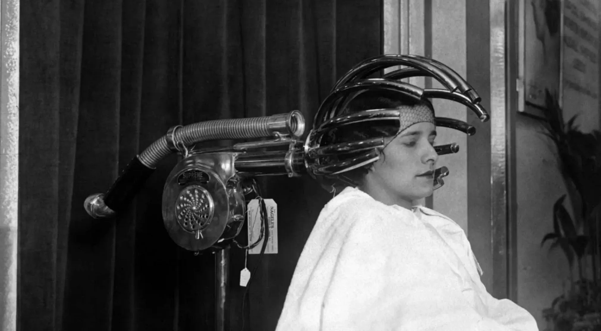 Black and white photo of a woman getting her hair permed with a vintage item. Guess the vintage item in this historical image.