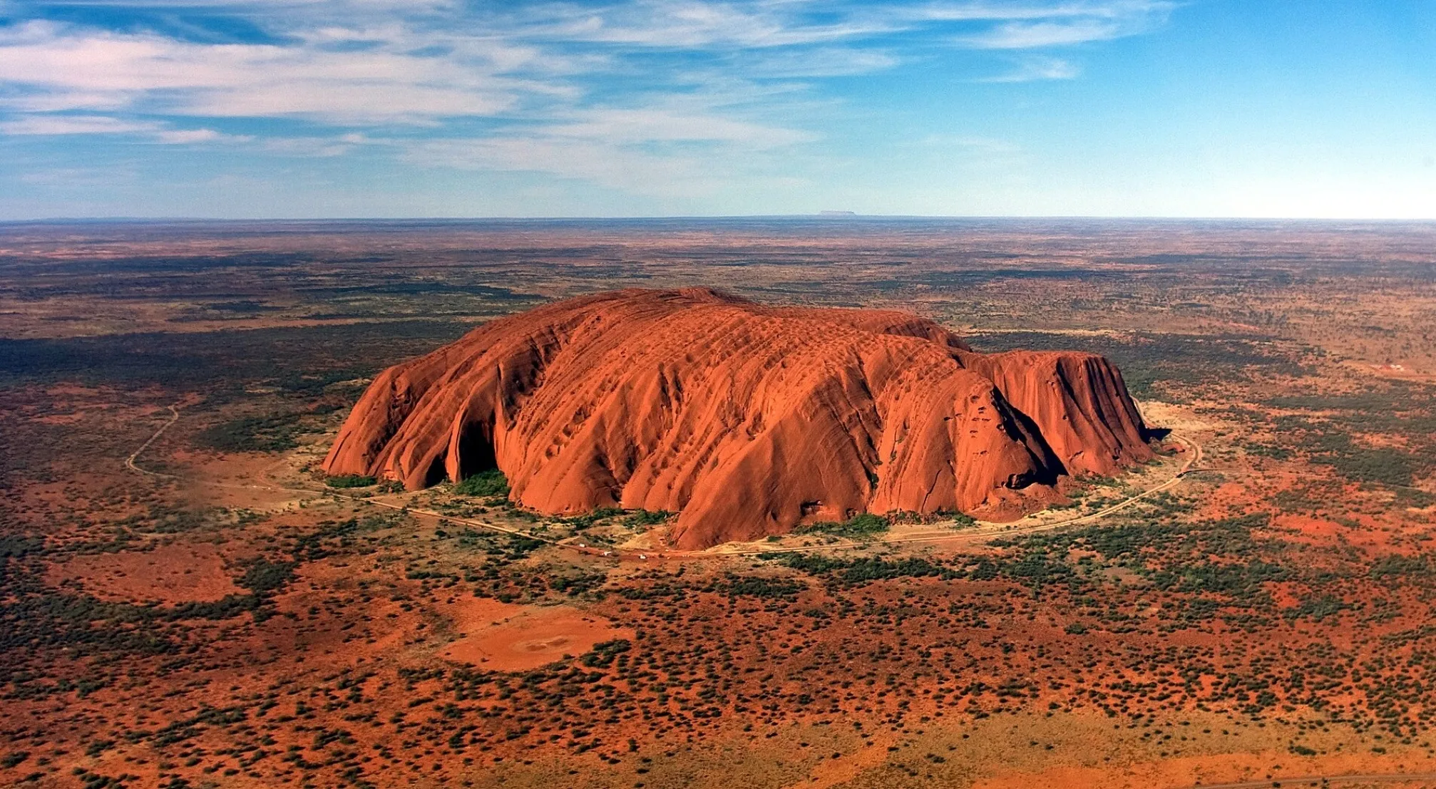 Aerial view of Uluru, a massive red geological structure in the Australian desert under a vast blue sky.