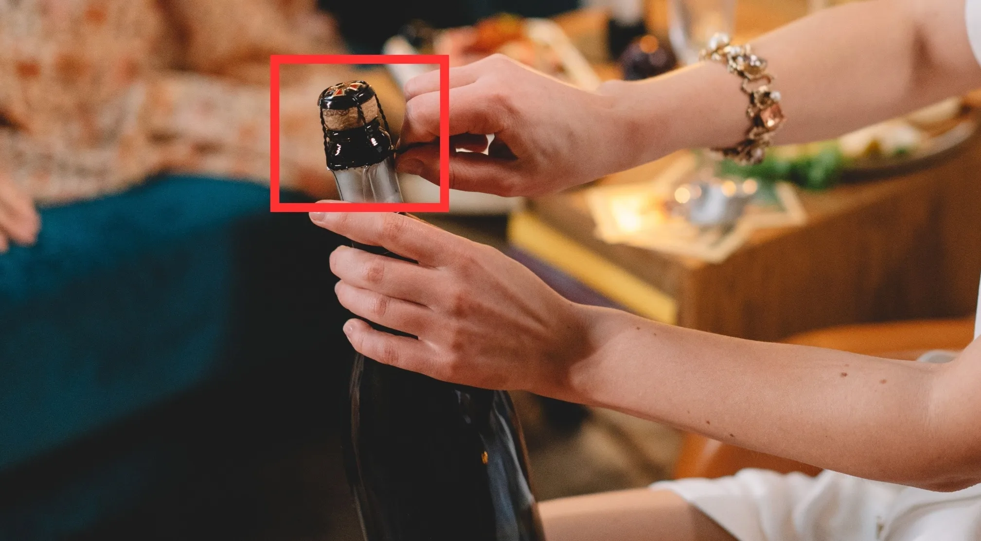 Hands removing the object from the top of the bottle, specifically a cork wrapped in dark foil, for a difficult word.