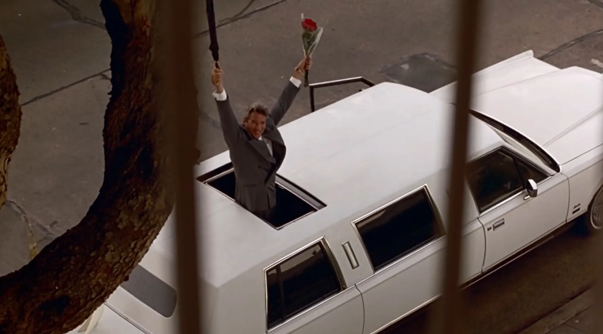 Man standing in a limousine sunroof holding an umbrella and a red rose, part of a movie's final scene.