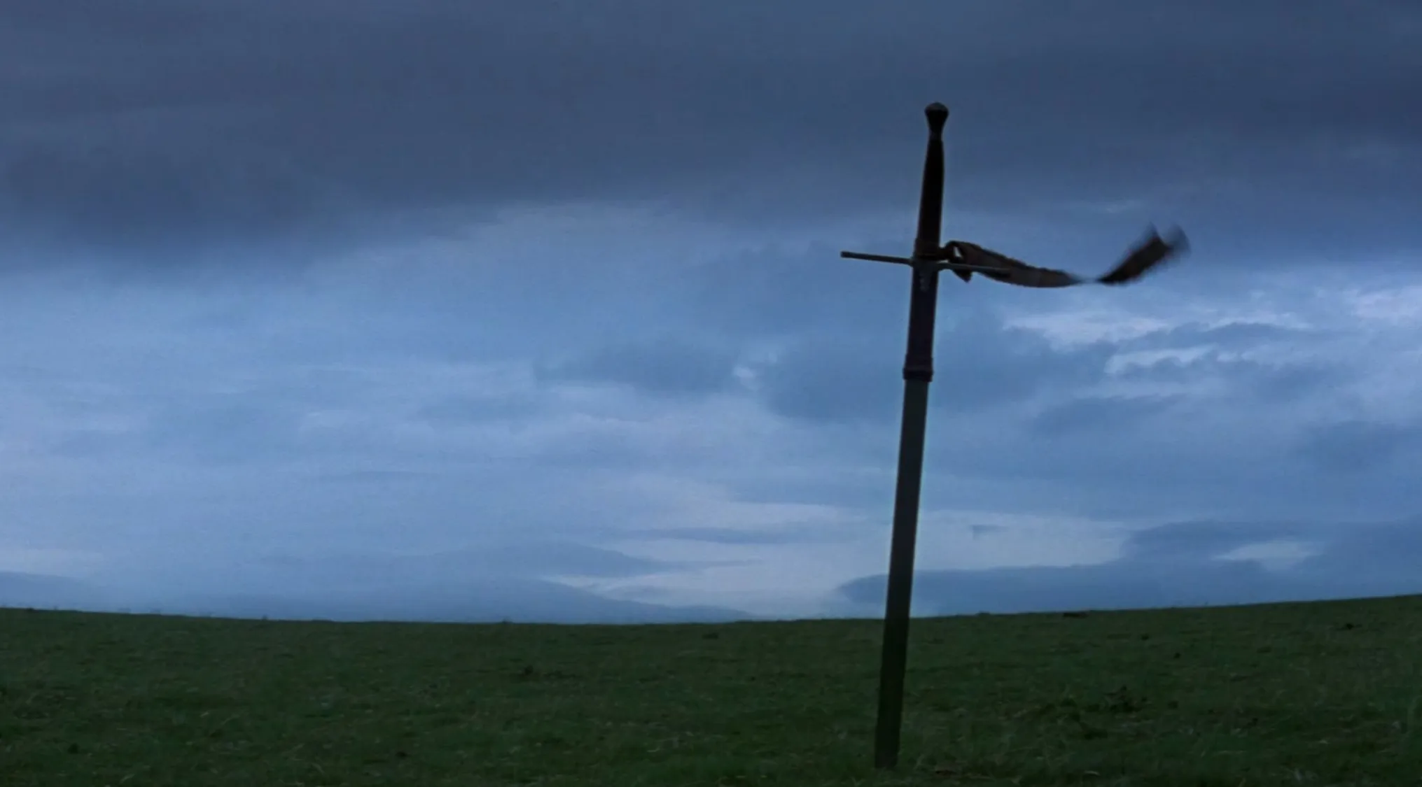 A sword planted in a grassy field under a cloudy sky, marking a film's final scene before the credits.