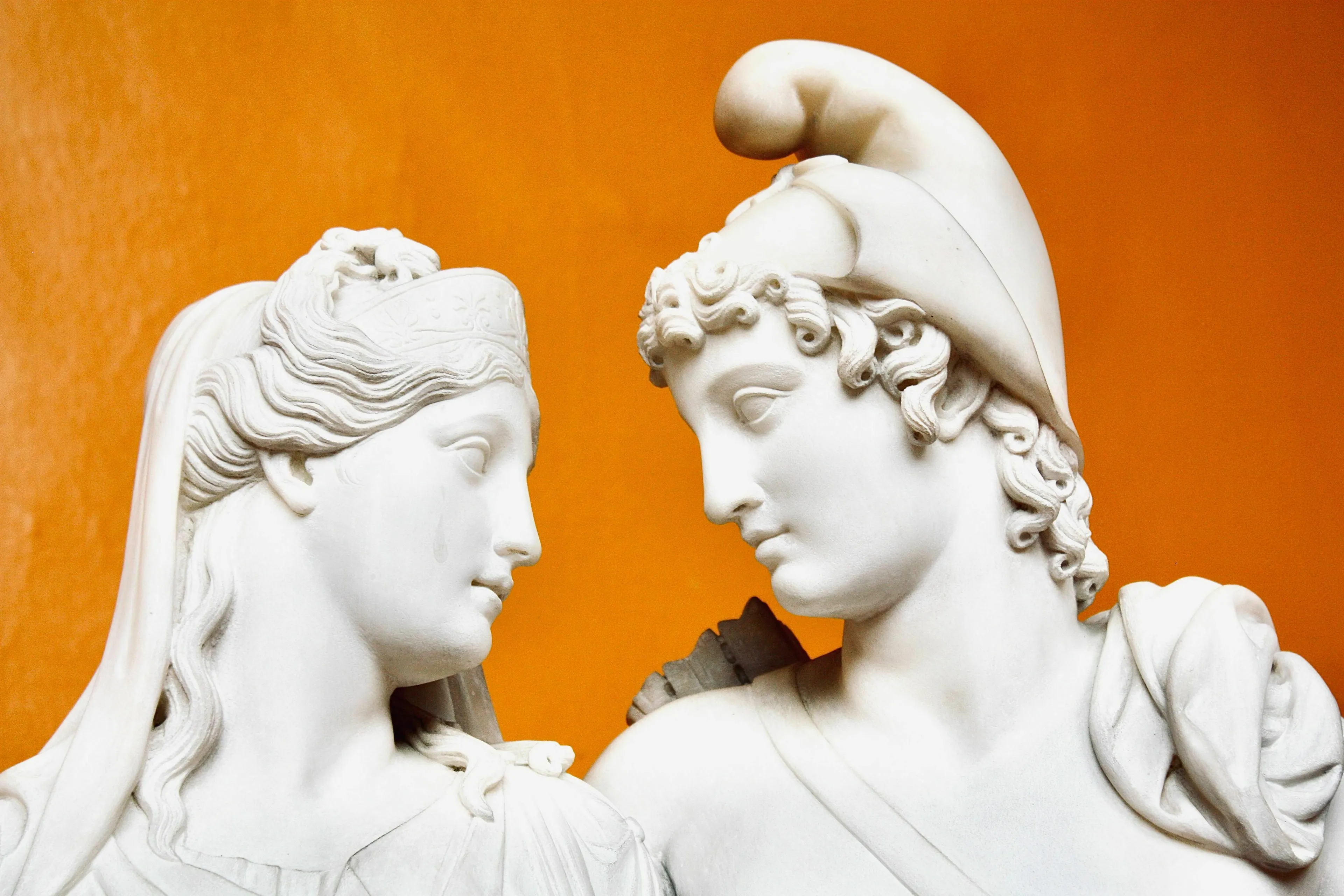 Two white marble statues, a male and a female figure, face each other, depicting mythological tricksters amidst arguing gods.