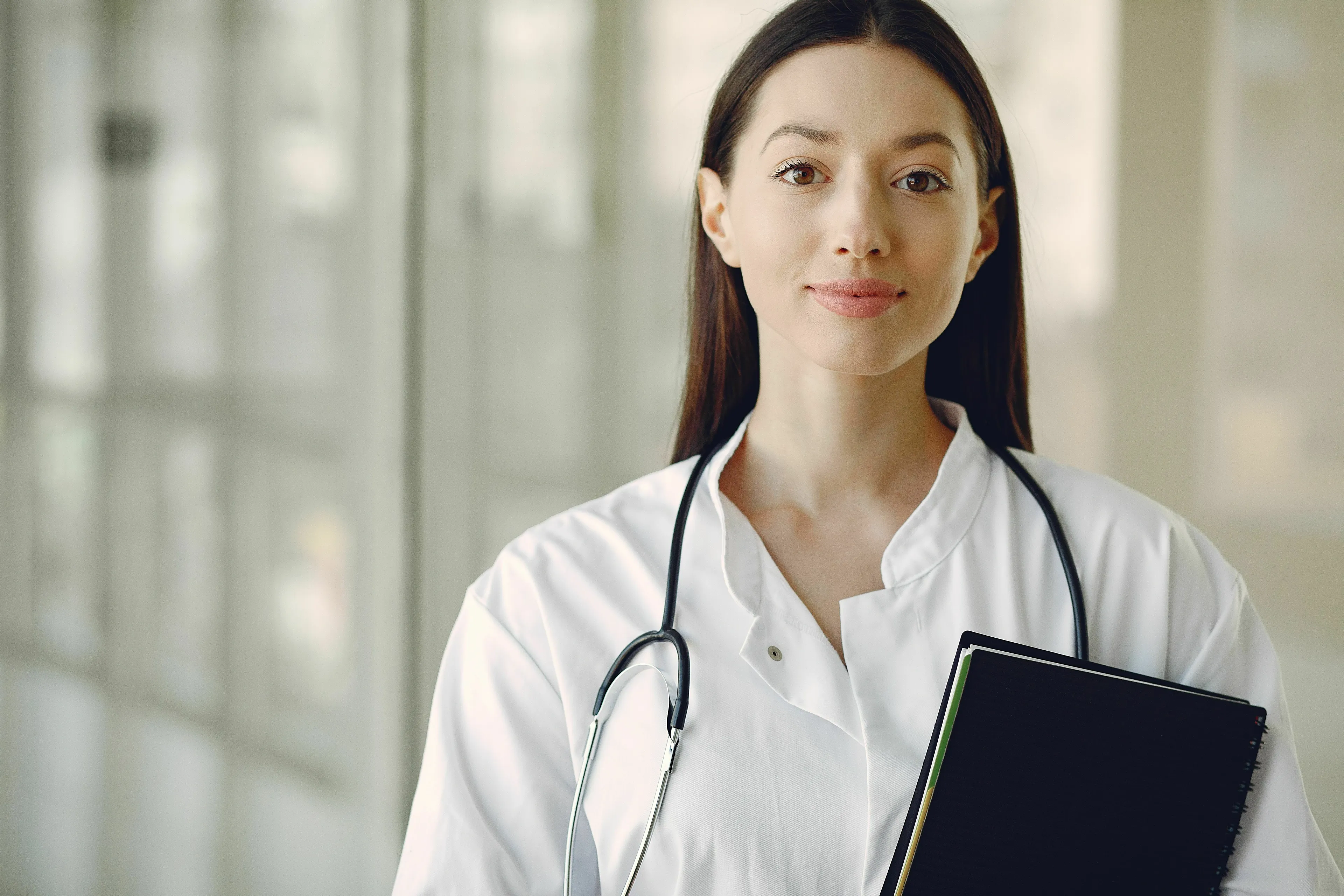 A female doctor in a white coat with a stethoscope, holding a folder, standing in a medical facility. A general knowledge quiz.
