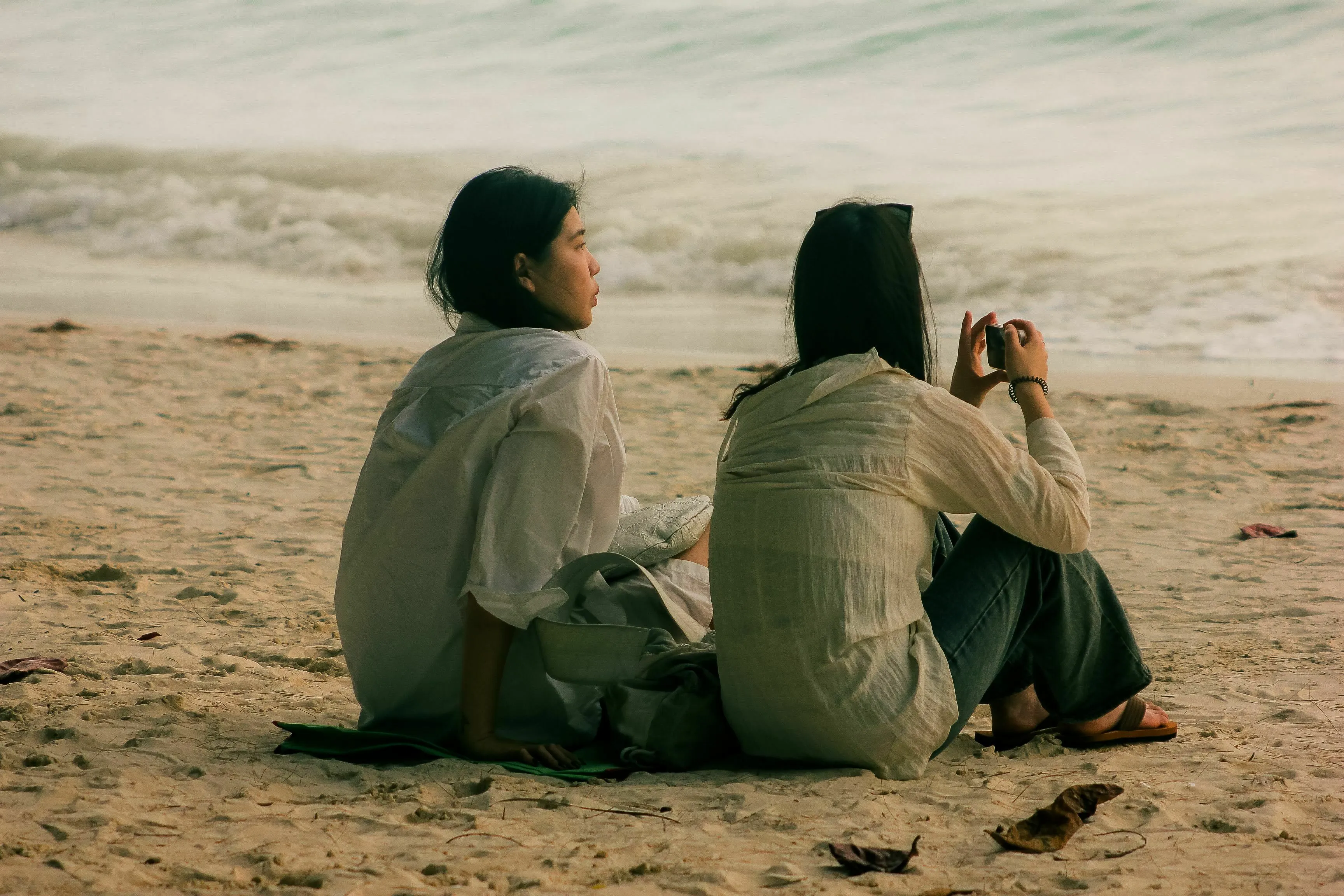 Two friends sitting on a beach, one looking out at the ocean and the other taking a photo, discussing a friend seeing a certain Angel Number.