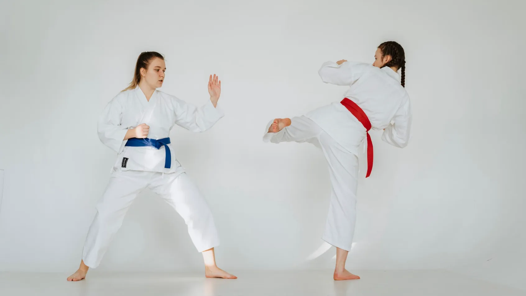 Two women in martial arts uniforms, one with a blue belt and one with a red belt, practicing kicks.