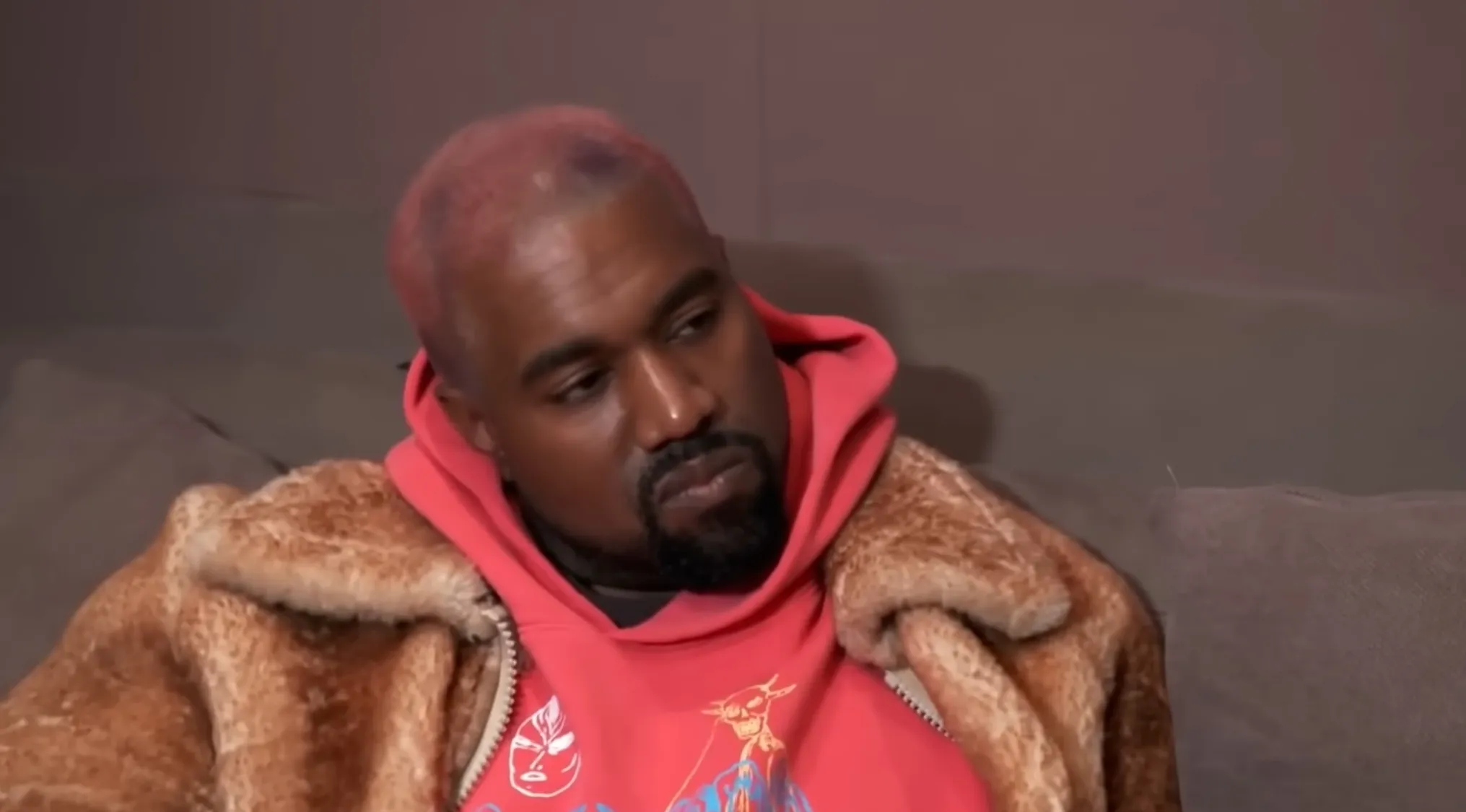 Kanye West, known for his appearances in reality TV shows, wearing a pink hoodie and fur coat, looking to the side.