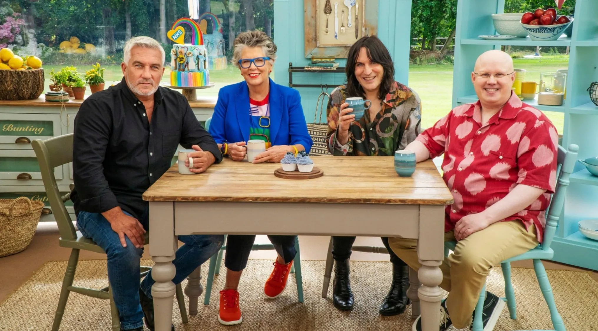 The Great British Bake Off hosts and judges, Paul, Prue, Noel, and Matt, seated at a rustic table.