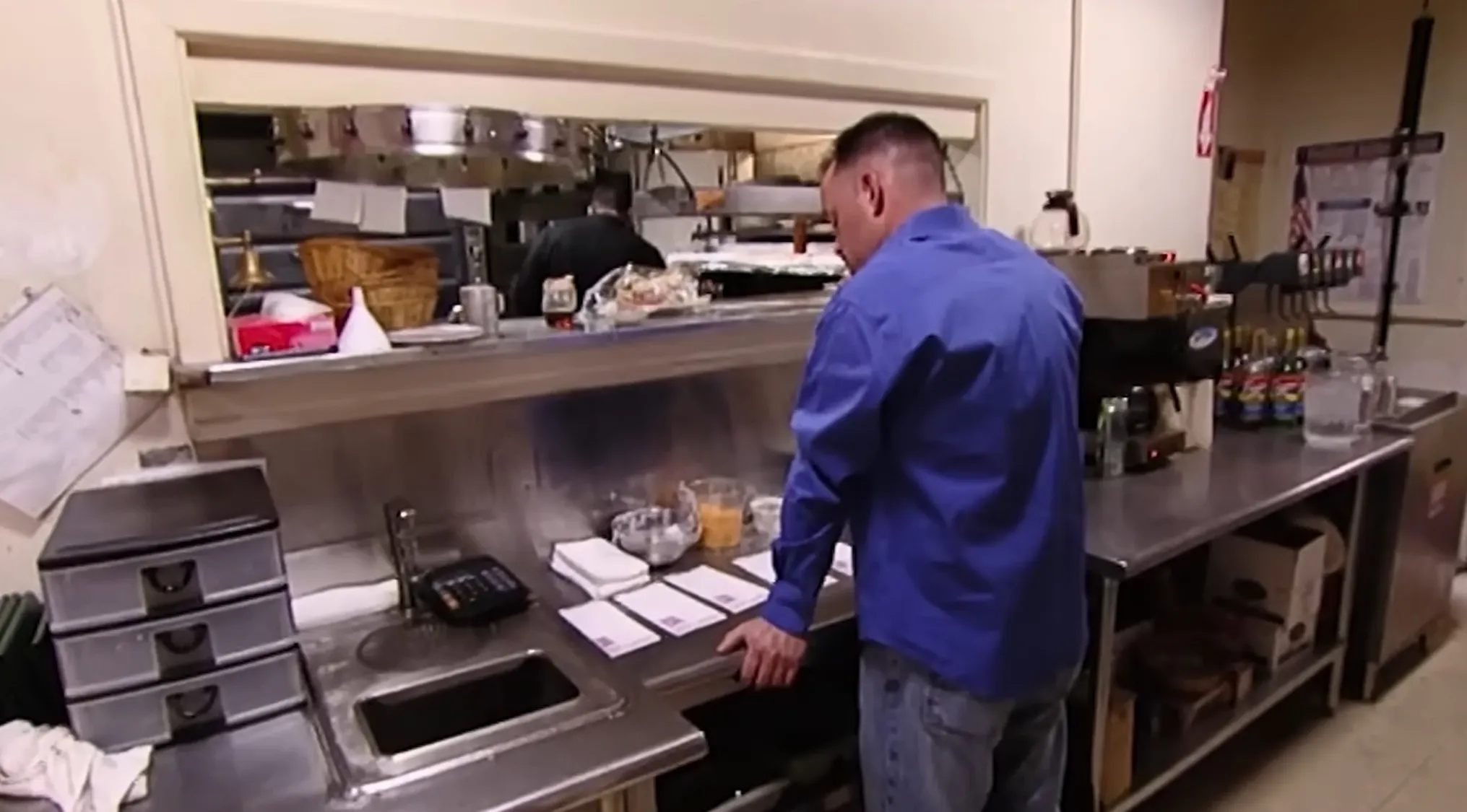 A chef, personality behind Kitchen Nightmares, looks at the items on a stainless steel counter in a commercial kitchen.