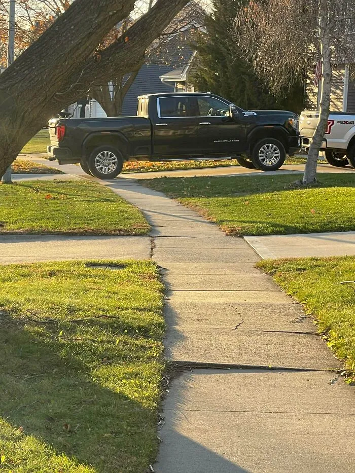 A large black pickup truck, showcasing bad parking, is parked on the grass blocking a public sidewalk.