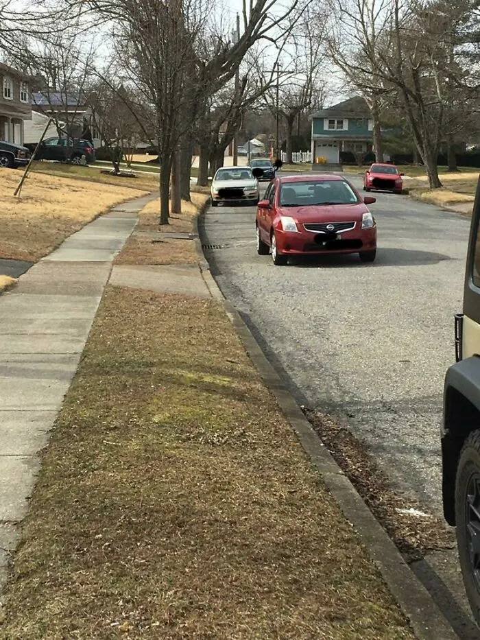 A red car, a prime example of jerk neighbors parking, is parked in the street partially blocking the flow of traffic.