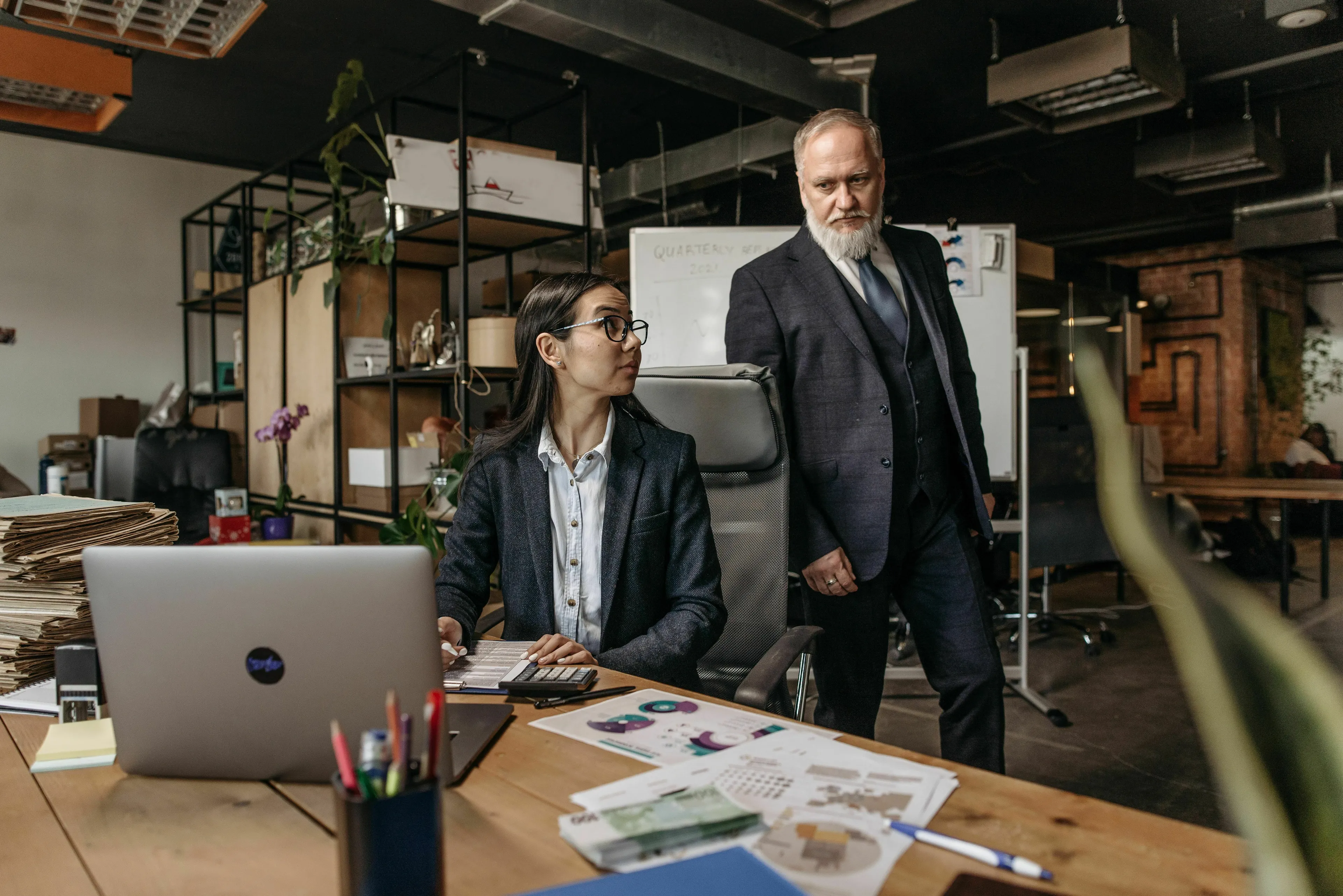 A boss observing an employee at a desk with a laptop and documents, illustrating what kind of liar you are regarding reports.