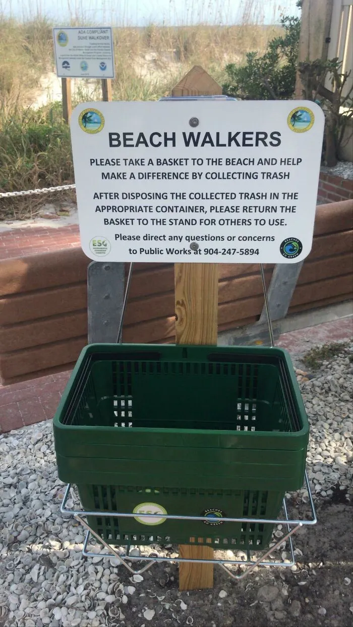 A sign for beach walkers to collect trash, with a green basket below it, promoting efforts to combat trash.