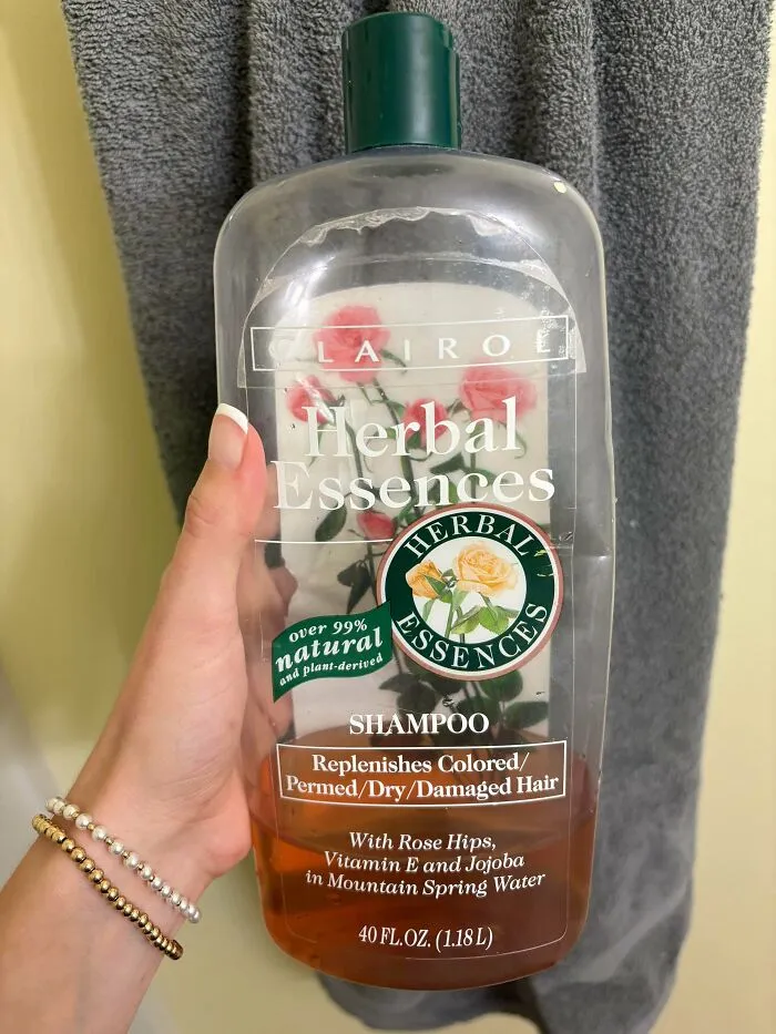A hand holding old products: a large, partially used bottle of Herbal Essences shampoo.