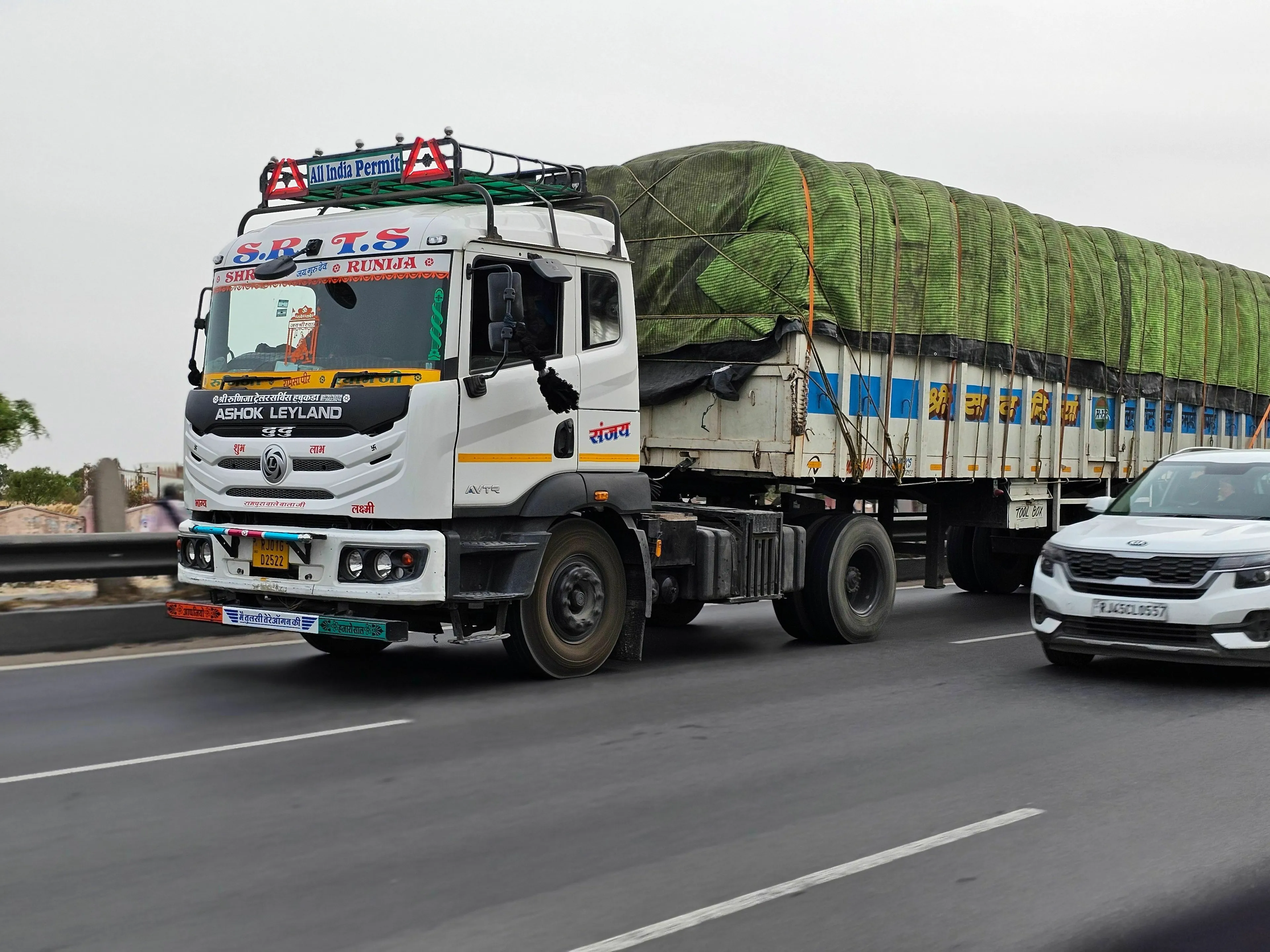 A large white truck with a green tarp covering its cargo, driving on a highway alongside a white car. American vs British English words.