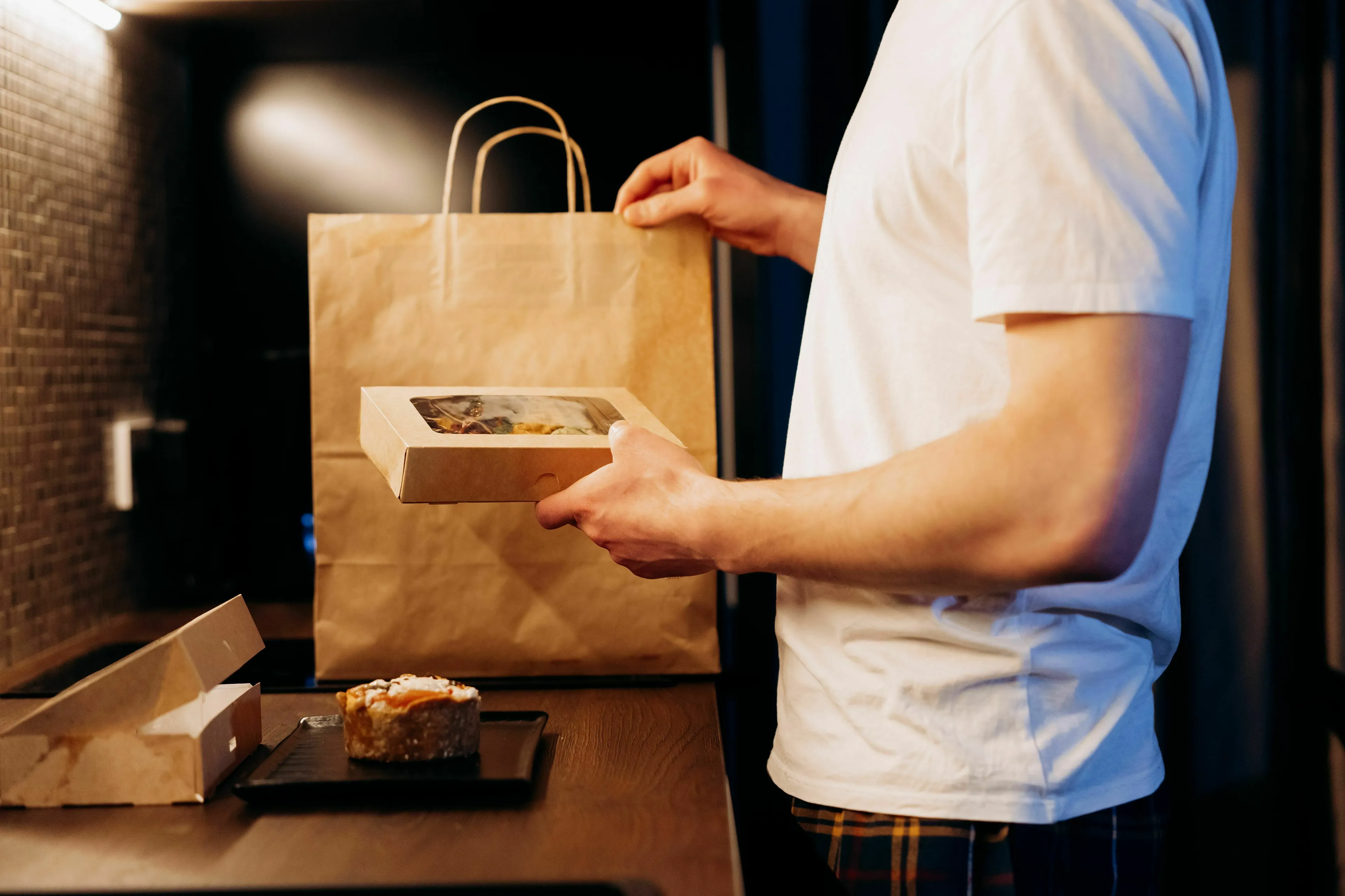 A person in a white T-shirt holding takeout food in a paper bag and a box. American vs British English words.