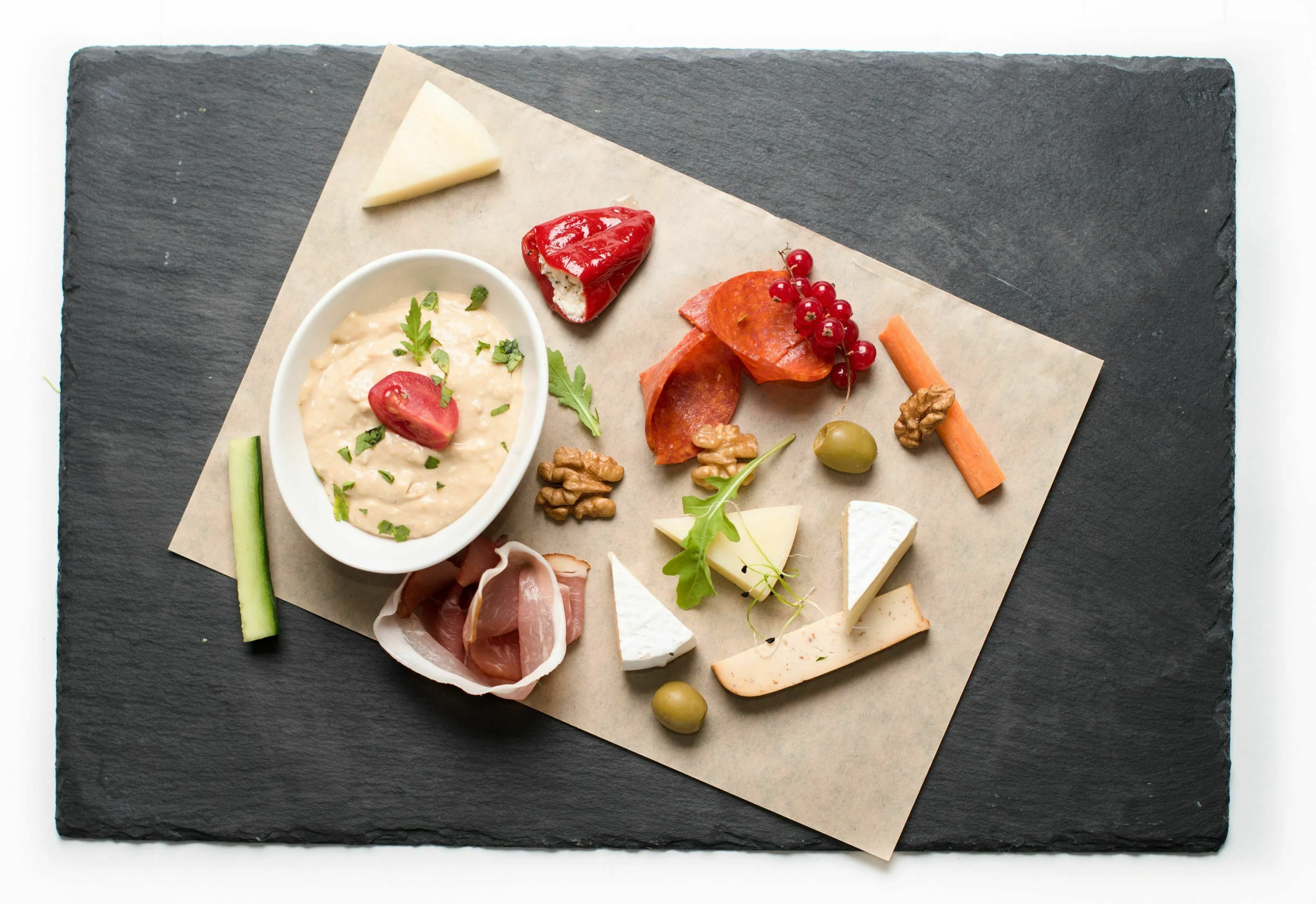 A delicious first course of a meal with various cheeses, meats, and a dip on a slate platter.