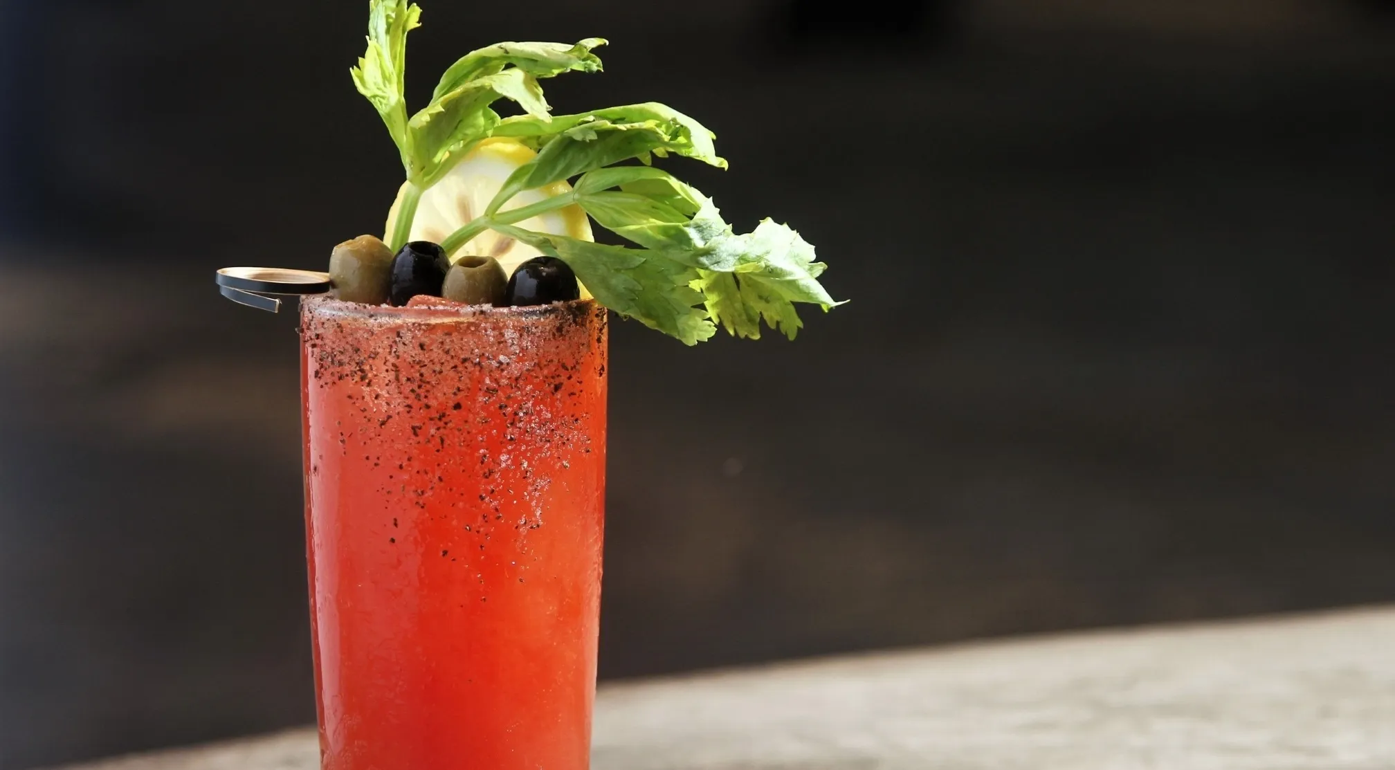 A Bloody Mary cocktail, garnished with celery, olives, and a lemon wedge, against a dark background, highlighting weird history facts.
