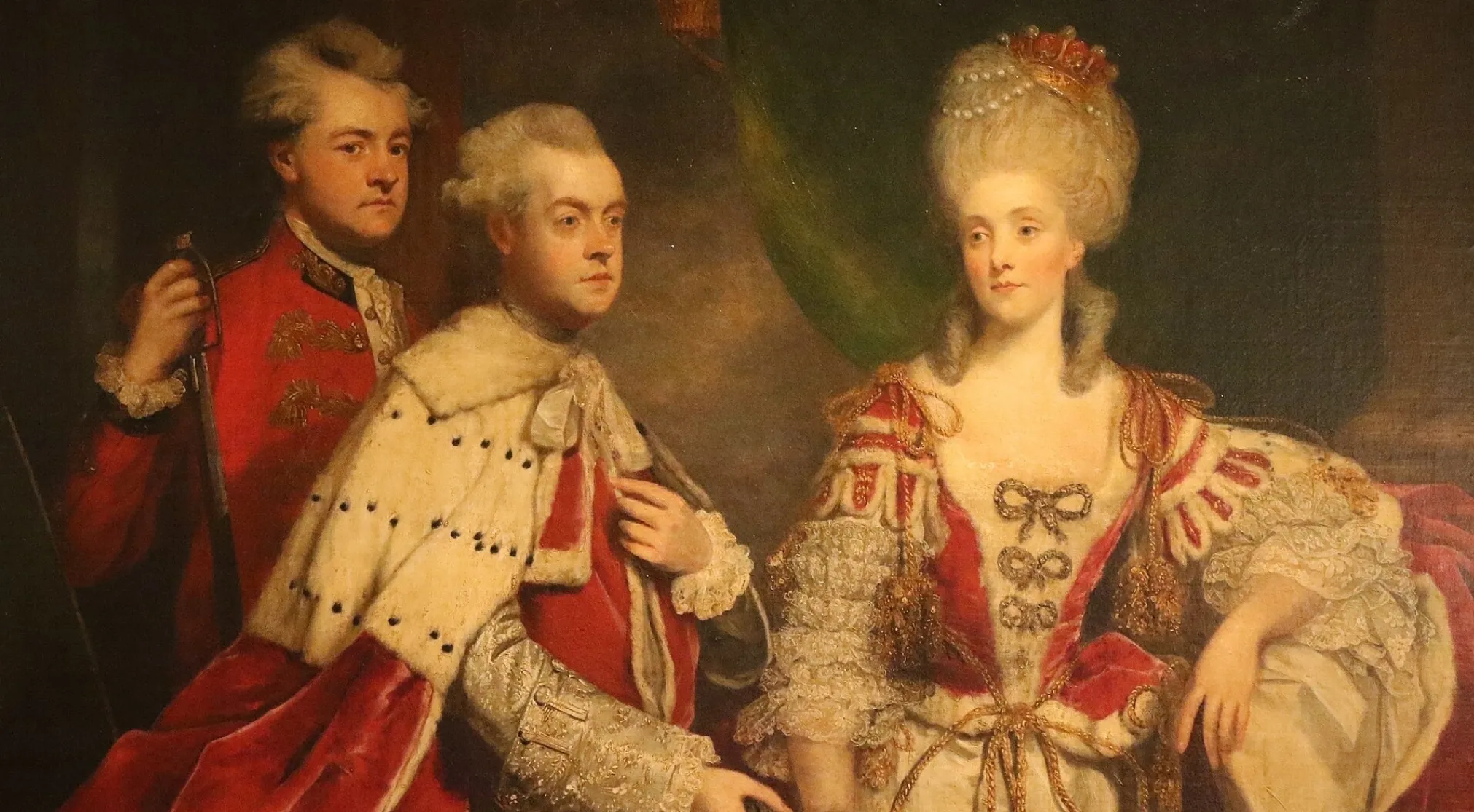 18th-century England royal painting. A man in red military attire, a man in a fur-trimmed robe, and a lady with a tall powdered wig.