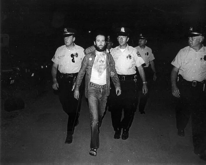 An impactful photo from history shows a bearded man in casual clothes being escorted by several police officers at night.