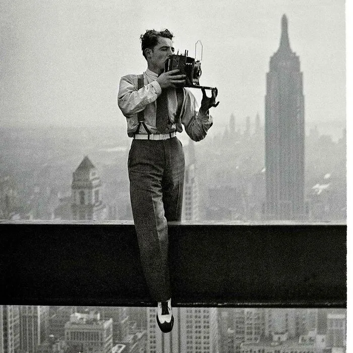 A photographer on a skyscraper ledge capturing a photo, an interesting and beautiful moment from history.