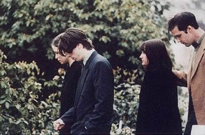 A poignant black and white photo from history features a group of individuals, appearing somber, walking outdoors surrounded by foliage.