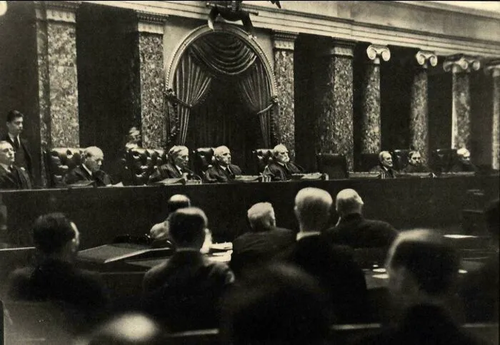 A black and white photo from history shows men seated in a grand hall, possibly a legislative body, with ornate architecture.