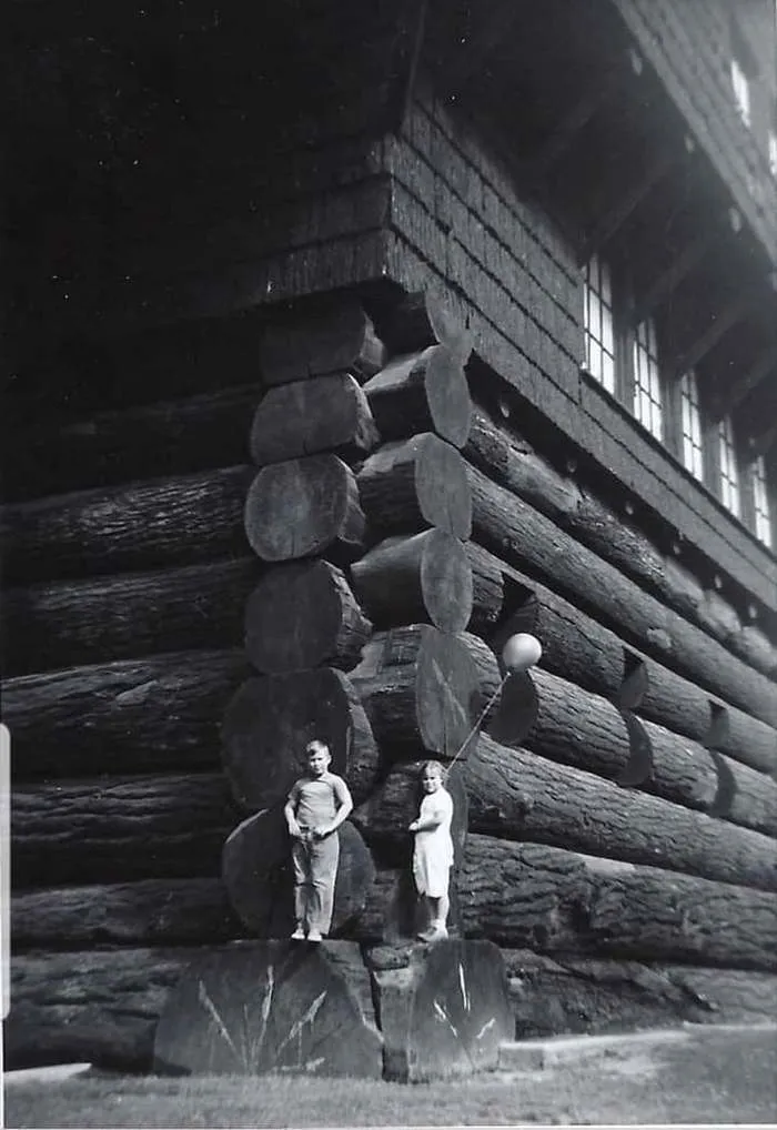 A heartbreaking black and white photo from history showing two children standing on large logs at the corner of a rustic building.