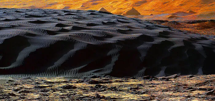 A breathtaking landscape revealing fascinating finds: dark, rippled sand dunes under an orange sky with distant peaks.