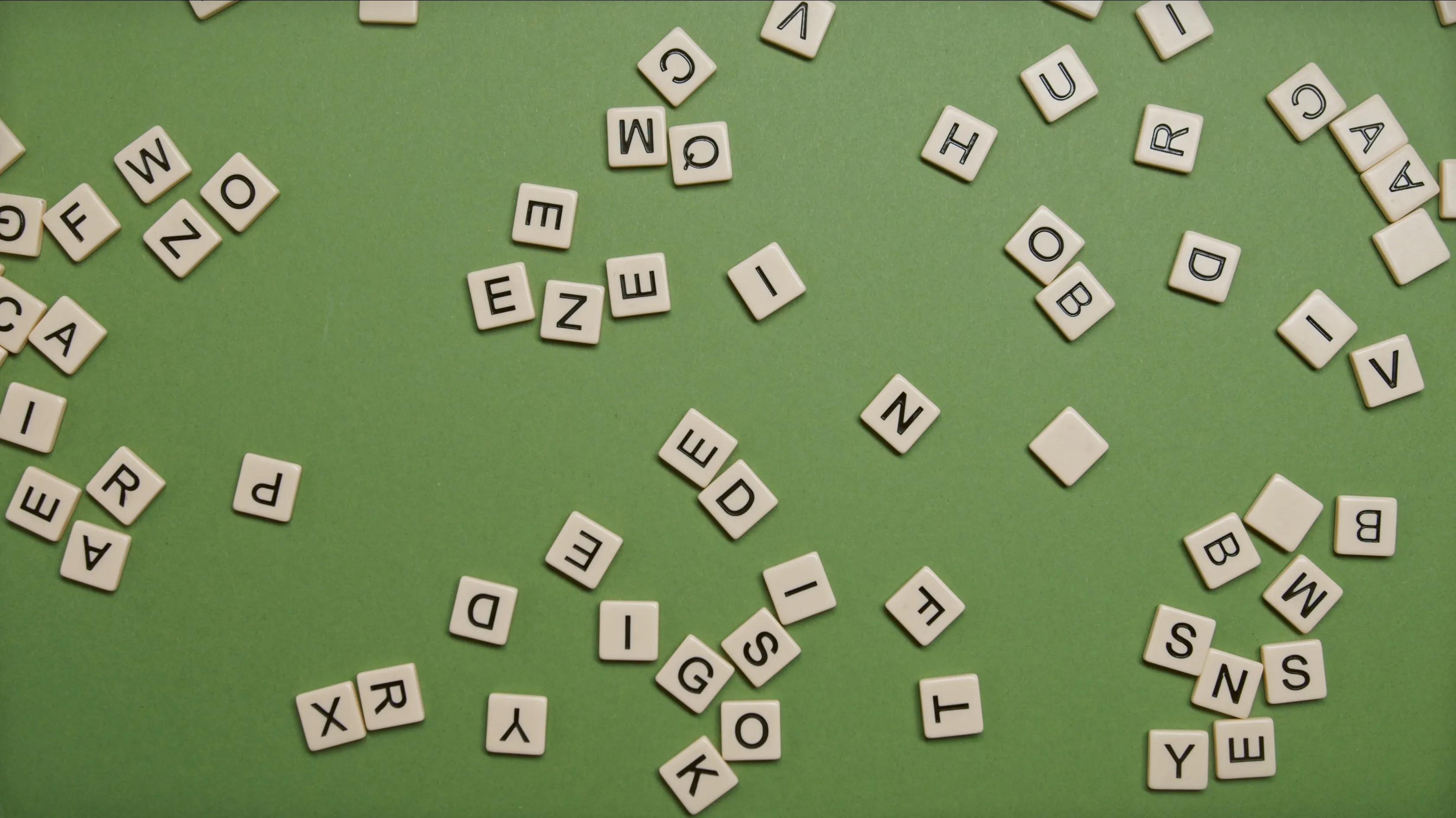 An overhead view of numerous English letter tiles scattered across a green surface, demonstrating a word puzzle.