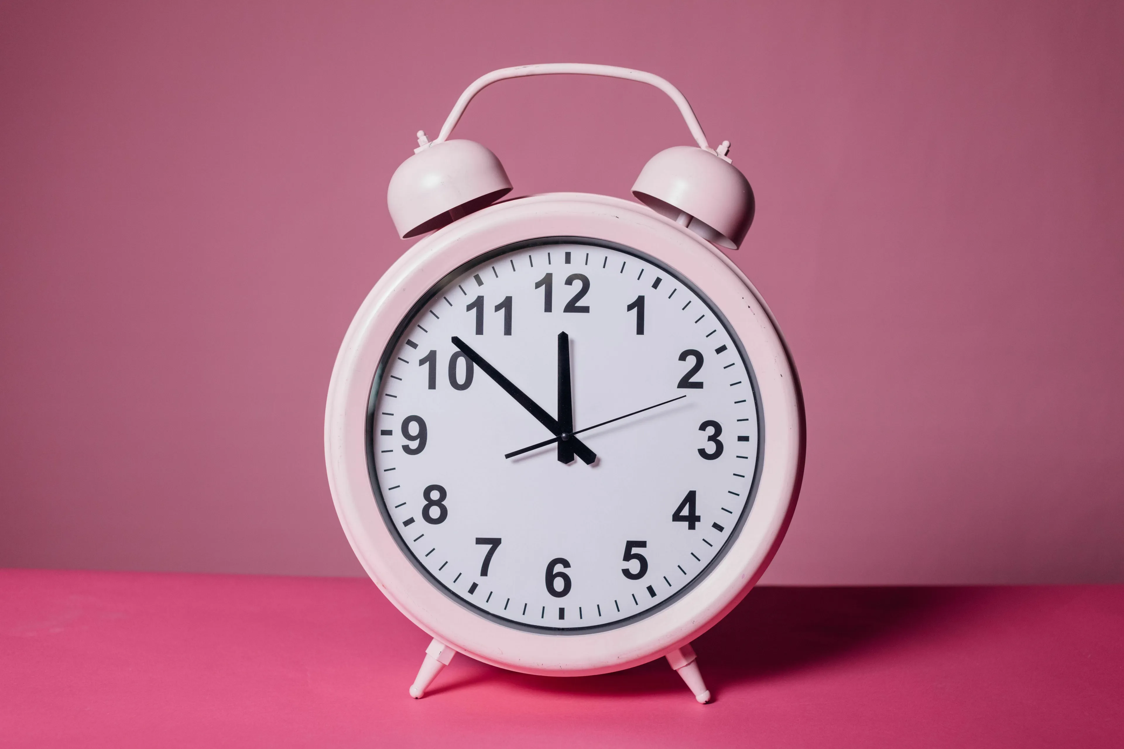 A pink alarm clock on a pink background, with the hands pointing to just before 12 o'clock, illustrating the math question.