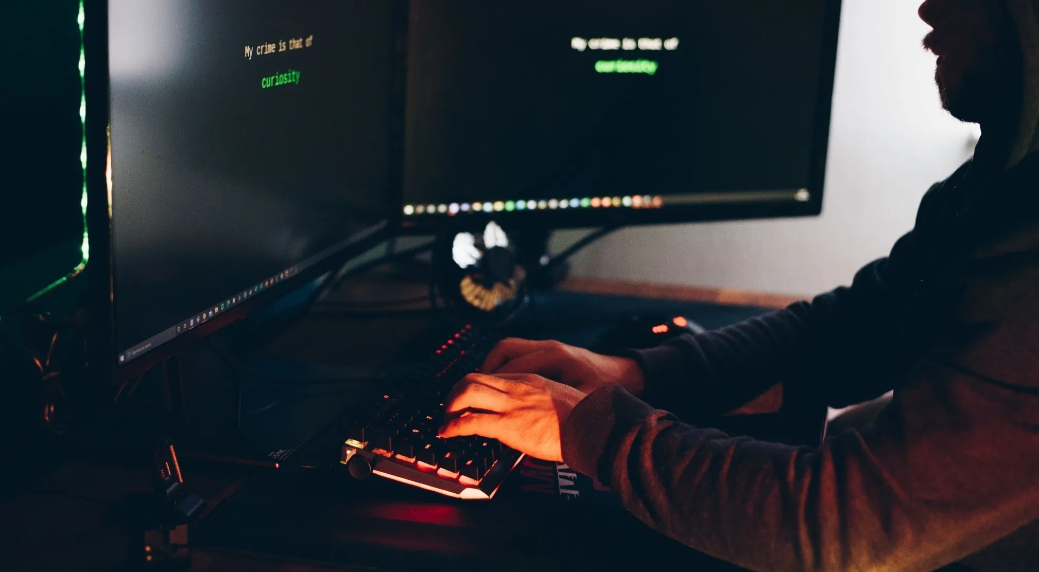 Person typing on a keyboard in a dark room with two monitors displaying text. Addresses malware and defective software.